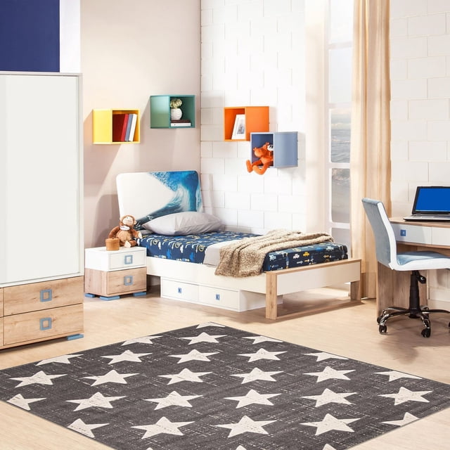 ECARPET Kids Room, Nursery, Playroom, Bedroom Area Rug, Angel Collection showing a grey area rug with white stars on a light wood floor in a child's bedroom. The room features a single bed with a blue and white wave headboard, bedding with vehicle prints, a beige throw, and a grey pillow. Colorful wall-mounted cube shelves in yellow, green, orange, and blue hold toys and books. A white nightstand and wardrobe with blue square handles, plus a desk with a blue office chair, complete the space. The rug is made from ultra-soft yarn for durability and softness, suitable for high-traffic kids' rooms and pet-friendly homes.