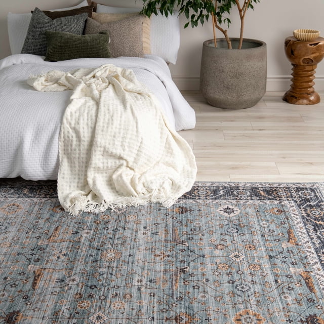Rug Branch Emir Collection Tapis Oriental Traditionnel Hydrofuge Présentant un Motif floral Bleu et beige Usé avec des Bordures Complexes, Posé sur un Plancher de Bois Franc Clair dans une Chambre avec une Literie Texturée Blanche, des Coussins Neutres, une Couverture Jetée Crème, un Grand Pot Gris avec une Plante Feuillue, et une table d'Appoint en Bois Sculptée, Idéal pour les Espaces Intérieurs à Fort Passage.