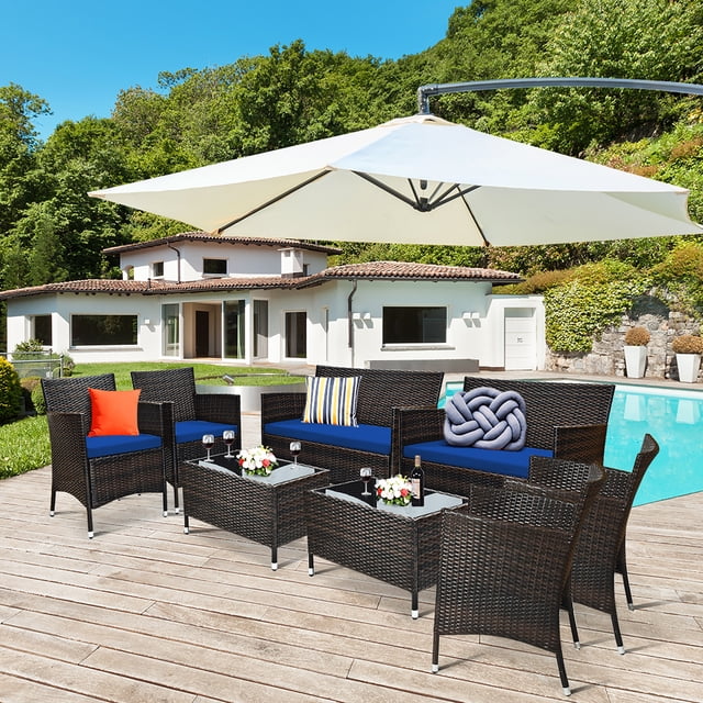 Costway 8PCS Rattan Patio Furniture Set Cushioned Sofa Chair Coffee Table Navy displayed in an outdoor setting by a poolside. The set includes four single chairs, two loveseats, and two coffee tables with tempered glass tops. The navy cushions provide a vibrant contrast to the dark rattan weave. The furniture is arranged on a wooden deck under a large white umbrella, with a modern house and lush greenery in the background.