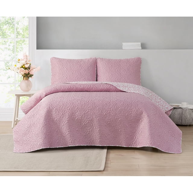 Valerie 3 Piece Reversible Quilt Set in muted pink with stitched large floral outlines on soft matte satin front and a white floral botanical pattern on the microfiber reverse, displayed on a bed with two matching shams in a bright bedroom with light wood floors and a beige rug. The machine-washable set adds warmth and subtle botanical style to bedroom decor.