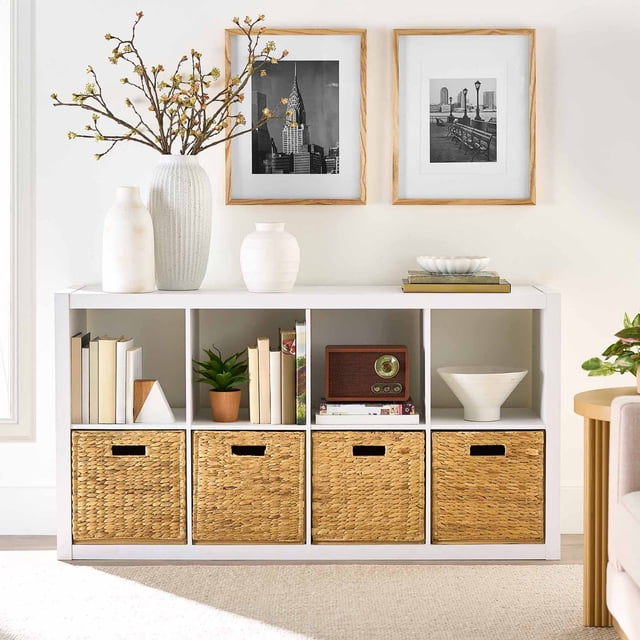 Better Homes & Gardens Organiseur de Rangement à 8 Cubes Bibliothèque Blanche à Cubes avec Huit Compartiments de 13 Pouces x 15 Pouces x 13 Pouces, Quatre avec Bacs de Rangement Tressés. la Rangée Supérieure Tient des Livres, Plante en Pot, radio vintage et Bols Décoratifs. Présente un Dos Ouvert pour la Gestion des Cordons, Conçue avec des vases Blancs et des photos Encadrées en Noir et Blanc dans un Salon Lumineux.