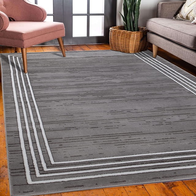 JV Home Tapis de Couloir Chic Carpet Tapis Coureur Ou Paillasson/tapis en Couleur Grise avec un design Moderne à Bordure Rayée. le Tapis Est Exposé dans un Décor de Salon avec un Plancher en Bois, un Fauteuil Rose, un Panier en Osier et un sofa Gris. Fabriqué à Partir de 100 % de Polypropylène avec un Endos en jute, Il Présente une Hauteur de Poil de 0,4 Po pour le Confort et la Durabilité. Offert en Plusieurs Tailles, Incluant 2x3, 2x7, 3x5, 4x6, 5x7, 5x8, 7x9 et 8x10, Convient aux Salons