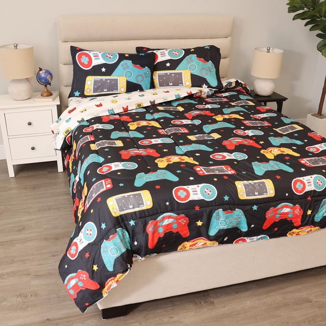 5-Piece Full/Double Bedding Set - Gamer with a reversible comforter featuring colorful video game controllers in red, yellow, teal, and white on black, including matching pillowcases and sheets with gaming designs, displayed on a beige upholstered bed in a kids' room with wooden flooring and bedside lamps.