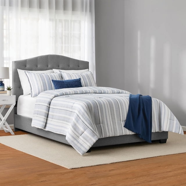 Mainstays 5-Piece Yarn Dyed Stripe Comforter Set, King on a grey tufted upholstered bed in a bright bedroom. The set features blue and white woven stripes on gauze fabric, including a comforter, two king shams, a chenille decorative pillow, and a navy blue fringe throw draped at the foot. The room has light grey walls, sheer white curtains, a white nightstand with a lamp and plant, and a beige rug on wood flooring. Suitable for year-round comfort and easy machine care.