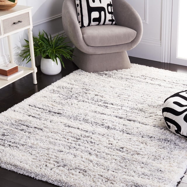 SAFAVIEH Fontana Dalia Distressed Stripes Plush Shag Area Rug with 2-inch plush pile in white and grey tonal stripes, placed on dark hardwood floors beside a grey modern chair with a black and white geometric cushion, a matching floor pillow, a white side table, and a green potted plant, ideal for adding comfort and style to living spaces.