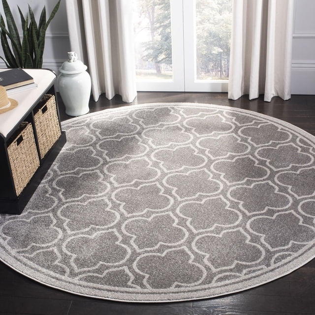 Safavieh Amherst Wilson Geometric Area Rug, round with a grey and white geometric trellis pattern, power-loomed polypropylene construction for durability and easy care, suitable for high-traffic indoor and outdoor areas, displayed on dark hardwood flooring near a window with white curtains and a black storage bench with wicker baskets.