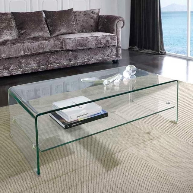 Heavenly Collection Waterfall Glass Table with Top Shelf, clear tempered glass coffee table with a modern waterfall design and open lower shelf holding books, placed on a beige textured rug in a living room with a grey velvet sofa and large window overlooking water, ideal for modern living rooms or offices.