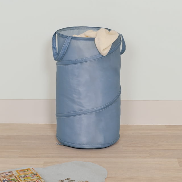 Neatfreak Kids Pop-Up Hamper in blue with collapsible cylindrical design and easy-carry handles, shown upright with a cream cloth inside on light wood flooring against a pale wall in a child's room, suitable for storing laundry, toys, and encouraging tidiness.