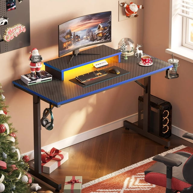 Bestier 42-inch gaming desk in blue with RGB LED lights, a monitor stand, headphone hook, and cup holder. The desk features a durable carbon fiber surface and adjustable foot pads for stability on uneven floors. It is shown in a festive room setup with a gaming PC, curved monitor, keyboard, and mouse on the desk, surrounded by holiday decorations, making it an ergonomic and stylish choice for gamers.