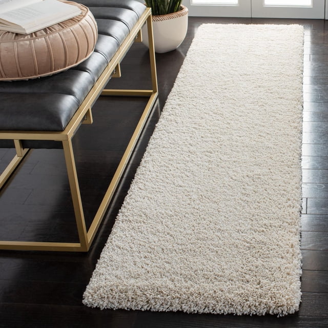 Safavieh Milan Harlow Tapis Shag Uni en Ivoire, Coureur en Fil Synthétique Tissé Mécaniquement à texture Épaisse et Douce, Posé sur un Plancher de Bois Franc Foncé à Côté d'Un Banc en Cuir Noir avec Cadre en Métal Doré et Coussin beige, Près de Plantes en Pot et de Portes en Verre, Convient pour Ajouter Chaleur et texture à la Déco Contemporaine de la Maison.