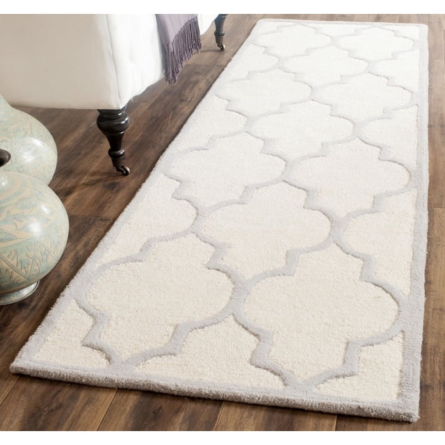 Safavieh Cambridge Kevin Geometric Area Rug with a cream background and raised light gray geometric trellis pattern, hand-tufted wool with plush cut and high/low loop piles, shown on a dark wood floor beside a white upholstered chair and decorative ceramic vases, ideal for adding texture and warmth to home or office decor.
