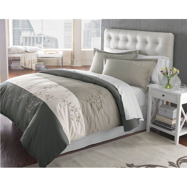 Springmaid Adelaide D/Q Duvet Cover Set on a bed with a white tufted leather headboard in a modern bedroom. The duvet cover features alternating grey and taupe panels with classic embroidered vine patterns on 100% cotton fabric. Matching pillow shams complete the set. The bed is styled with white sheets and a white nightstand holding a glass vase with flowers and books underneath, with a city skyline visible through large windows. The set is suitable for adding a contemporary look to bedroom decor.