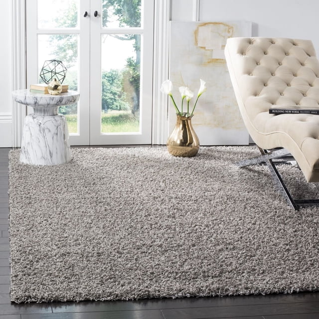 Safavieh Athens Tapis Shag en Peluche Uni Gris Pâle avec un Poil Doux et dense en Polypropylène, Installé sur des Planchers de Bois Franc Sombres dans une Pièce Lumineuse Comprenant une chaise Longue Capitonnée beige avec Pattes en Métal, une table d'Appoint en Marbre Blanc avec Livres et Déco, et un vase Doré Tenant des Callas Blancs Près de Portes en Verre.