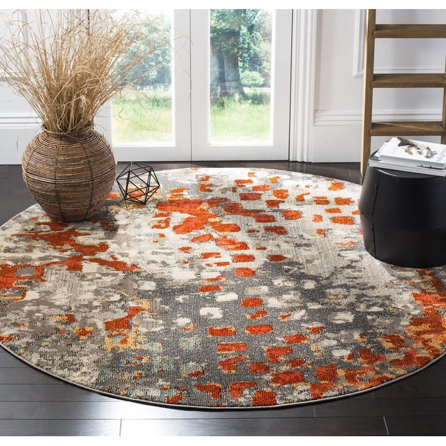 Safavieh Monaco Driskoll Abstract Area Rug, 5-foot round in grey and orange featuring a durable polypropylene blend with a power-loomed mix of high and low loop pile, displayed on dark hardwood floor near a wicker vase with dried grasses and a black side table with books in a sunlit room. This accent rug is suitable for adding Bohemian-chic flair to living spaces or as a stylish floor centerpiece.