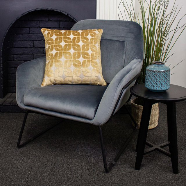 Haven Luxury Feather Filled Cushion with gold geometric pattern on a grey velvet armchair with black metal legs, beside a black round side table holding a blue textured ceramic vase, set near a dark brick fireplace and tall green grass in a woven basket on grey carpet, suitable for decorative and comfort use.