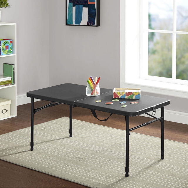 Mainstays Adjustable Height Table in classic black with fold-in-half design and metal legs with rubber stoppers, placed on a beige rug in a bright room. This rectangular folding table features a smooth resin surface and built-in carry handle, holding craft markers and alphabet puzzle pieces. Suitable as a portable folding table for indoor crafts, gatherings, or activities.