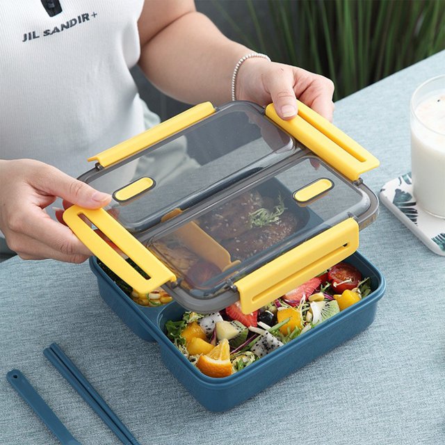 SuoKom Adult Lunch Box, 1100 ml 3-compartment bento lunch box in blue with yellow accents, designed for food storage. The box features a transparent lid with a silicone seal and four buckles for leakproof closure. It includes chopsticks and a spoon, and is microwaveable and dishwasher safe. Displayed on a table with fresh food inside, such as fruits and vegetables, making it ideal for home, office, school, or outdoor use.