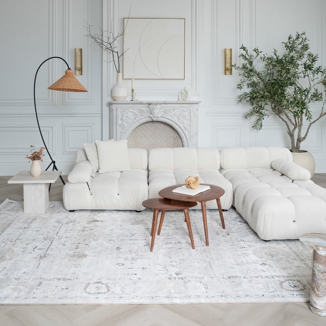Rug Branch Atlas Scandinavian Indoor Area Rug in beige ivory distressed pattern, 3'11" x 5'7", placed under a white tufted sectional sofa and two nested wooden coffee tables in a bright living room with white paneled walls, decorative white fireplace, and large potted green plant.