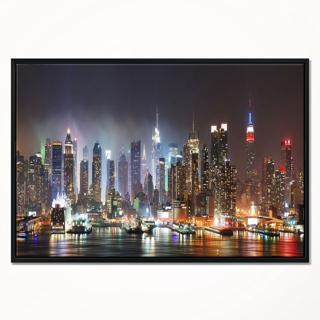 Design Art Lit Nyc Manhattan Skyline Framed Canvas Art Print showing a centered night view of Manhattan's illuminated skyline with reflections on the water, printed on fade-resistant canvas stretched over a 1-inch wood sub frame and gallery-wrapped in a black floating frame, includes hanging kit for easy wall mounting.