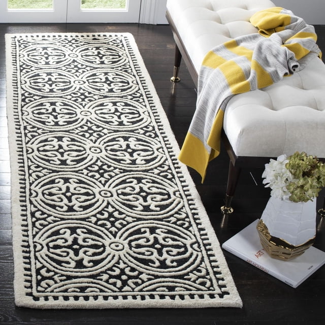 SAFAVIEH Cambridge Leslie Geometric Area Rug in black and white hand-tufted wool with circular medallion patterns, on dark hardwood floor beside a white tufted bench with a yellow and gray striped throw and a white vase with flowers on art books, suitable for modern home or office decor.