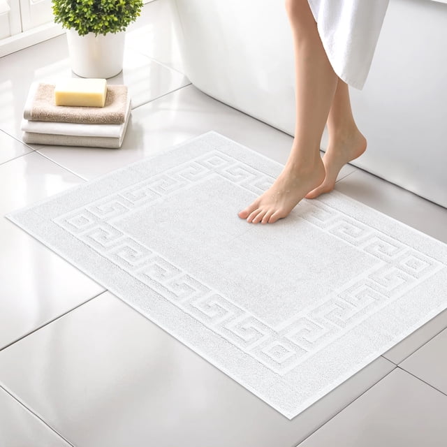 Canadian Linen Everest Bathmats, 2 Pack, 20"x30", soft absorbent cotton mats in pearl white with a Greek key design. These lightweight, quick-dry bathroom tub mats are shown in a modern bathroom setting with a person stepping onto one. The mats feature a smooth terry texture and are double-stitched for durability.