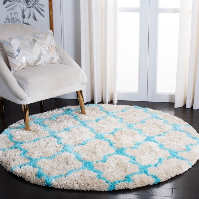 Safavieh Barcelona Anabelle Geometric Shag Area Rug, round with a plush beige pile and turquoise Moroccan quatrefoil pattern, placed on dark hardwood flooring beside a beige upholstered chair with wooden legs in a bright room with white curtains, ideal for adding texture and style to accent areas.