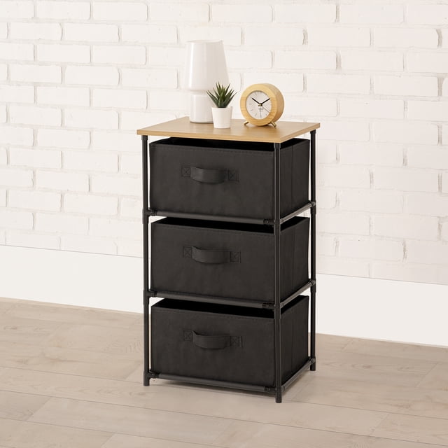 Mainstays Commode de Rangement à 3 Tiroirs avec Cadre en Métal Noir et Tablette Supérieure à Motif Grain de Bois, Dotée de Trois Tiroirs en Tissu Noir avec Poignées, Idéale pour Organiser Vêtements et Accessoires dans les Chambres Ou les Dortoirs, Présentée contre un Mur de Briques Blanc sur un Plancher de Bois Clair avec une Lampe Blanche, Petite Plante en Pot et Horloge Ronde en Bois sur le Dessus.