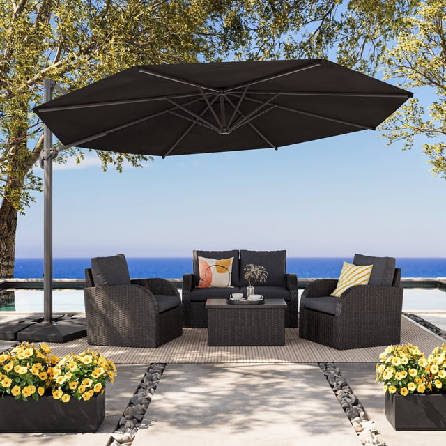 CorLiving 11.5ft Deluxe Tilting and Rotating Offset Patio Umbrella with aluminum frame and black durable canopy shading a dark wicker outdoor seating set with two armchairs, a loveseat with colorful cushions, and a coffee table on a beige rug by a pool with ocean view, featuring yellow flowers in black planters and surrounded by trees.