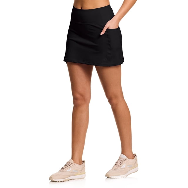 Reebok Women’s Ace High-waisted Skort with Pockets in black, front view on a model. Features a wide high-rise waistband, built-in inner shorts, and side pockets made from stretch Motion Poly/Span Interlock fabric. Suitable for tennis, golf, running, and casual athletic wear.