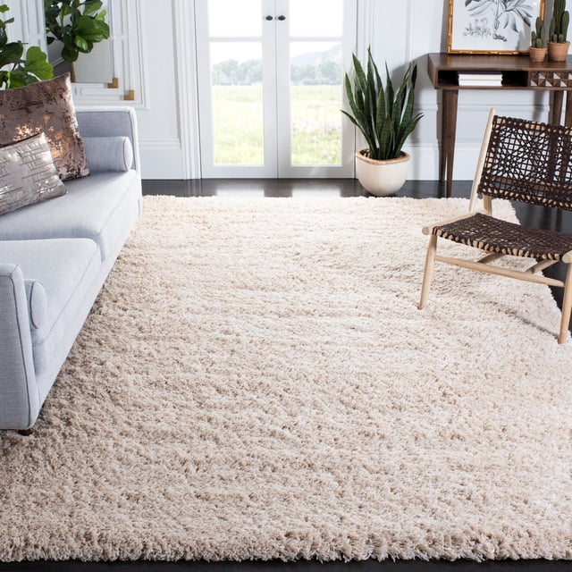 SAFAVIEH Polar Abbot Solid Extreme Plush Shag Area Rug with 3-inch thick plush pile in beige, placed on dark hardwood floors in a bright living room featuring a light grey sofa with metallic pillows, a woven dark brown chair, and green potted plants, ideal for adding warmth and texture to home decor.