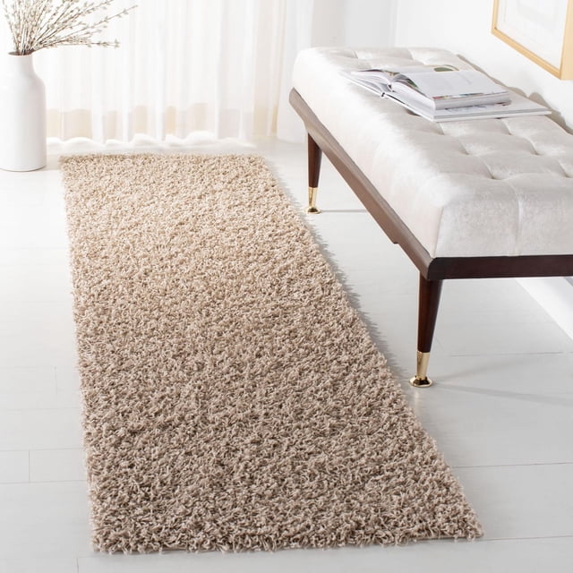 Safavieh Athens Solid Plush Shag Area Rug in beige on white hardwood floor next to a cream tufted bench with dark wood legs and brass feet, featuring a dense polypropylene shag pile that provides plush comfort and durability, suitable for transitional home decor and often used to add warmth to entryways or hallways.