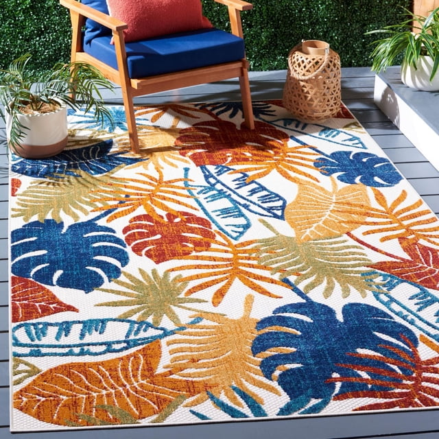 SAFAVIEH Cabana Kuzman Floral Outdoor Area Rug with a tropical leaf pattern in blue, rust, gold, and green on white, placed on a gray deck beneath a wooden chair with navy cushions and a woven lantern, made from durable synthetic yarns suitable for outdoor patios and porches.
