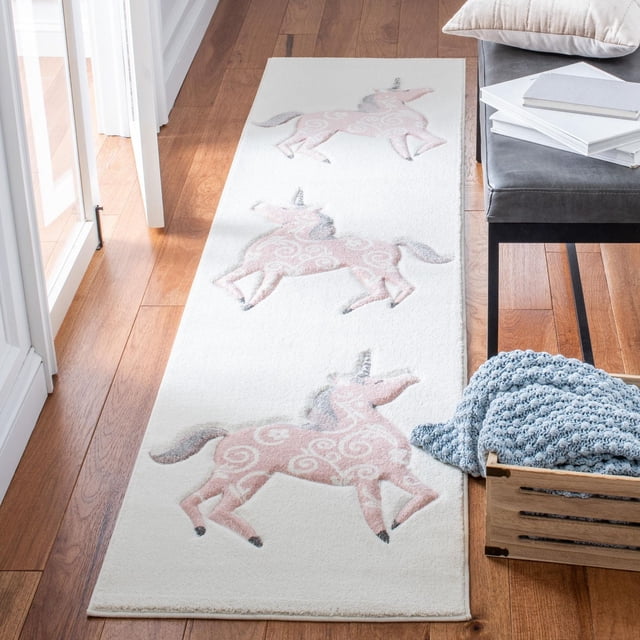 Safavieh Carousel Kids Dalach Unicorn Area Rug with a white background and three pink unicorns featuring swirled patterns, power-loomed from durable synthetic yarns, placed on a hardwood floor next to a gray bench and a wooden crate with a blue knitted blanket, suitable for kids decor and play areas.
