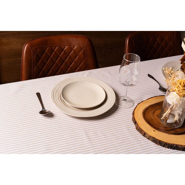 Fabstyles 100% Cotton Bistro Stripe Table Cloth in beige and white stripes on a dining table set with cream ceramic plates, a clear wine glass, silver spoon, and a wooden round slab centerpiece holding a glass vase with dried flowers, paired with brown quilted leather chairs. Suitable for indoor and outdoor dining and special events.