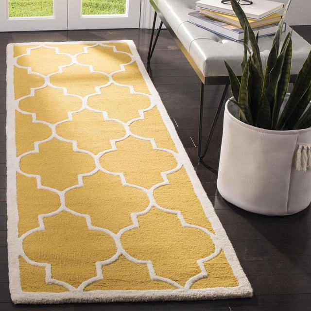 Safavieh Cambridge Kevin Geometric Area Rug in yellow with white geometric trellis pattern, hand-tufted wool featuring plush cut pile and texture-rich high/low loop and cut piles, placed on dark hardwood floor beside a white bench and potted snake plant near glass doors in a sunlit room, ideal for adding modern American style to home or office decor.
