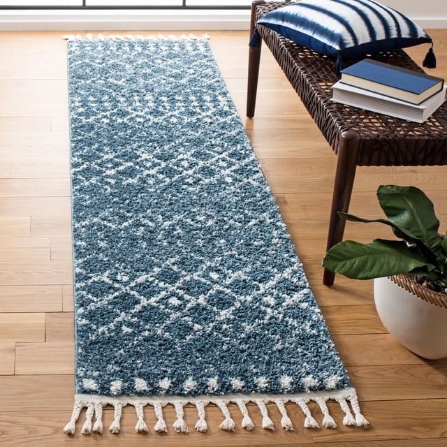 Safavieh Berber Fringe Robynne Tapis Shag Aztèque Bleu et Blanc avec une pile Peluche de 1,25 Po et des Motifs Aztèques Tribaux, Présentant des Franges Blanches aux Deux Extrémités, Placé sur un Plancher de Bois Franc Clair à Côté d'Un Banc Tressé avec un Oreiller Rayé Bleu et Blanc et des Livres, Accompagné d'Une Plante en Pot Verte. Ce Tapis de Style Bohème Convient Aussi pour Ajouter de la texture aux Couloirs Étroits Ou aux Entrées.