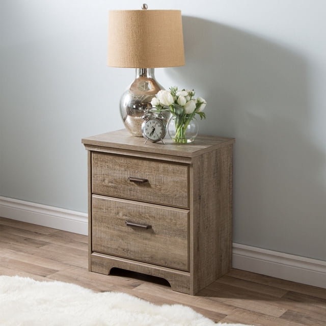 South Shore Versa 2-Drawer Nightstand in weathered oak finish with two metal-handled drawers and cutout kickplate base, shown with a beige fabric lamp, a small round alarm clock, and a glass vase of white tulips, suitable for bedside storage and organization.