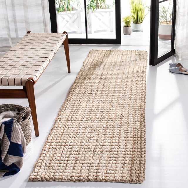 Safavieh Natural Fiber Aletha Solid Area Rug in natural beige woven sisal texture, displayed as a runner rug on white flooring beside a woven leather bench and a basket with a striped blanket, with sunlight from open glass doors and green potted plants outside, ideal for coastal or casual beach house style interiors.