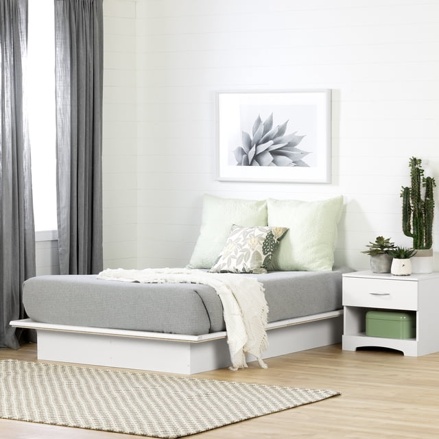 South Shore Step One Full Platform Bed in pure white with a low-profile solid base supporting a grey mattress without a box spring. The bed features clean lines and modern design, ideal for space-saving and discreet storage. It is styled in a minimalist bedroom with light wood flooring, grey curtains, a white nightstand with potted succulents, soft green and floral decorative pillows, and a black-and-white botanical print on the white shiplap wall.