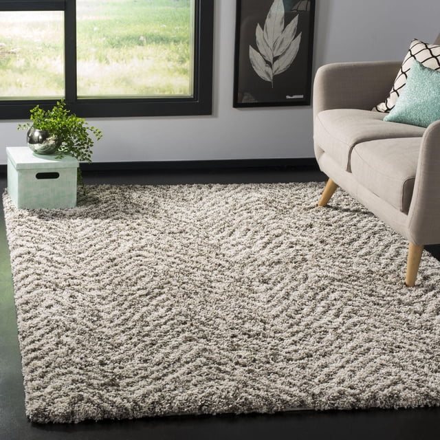 SAFAVIEH Hudson Adam Chevron Shag Area Rug featuring a plush synthetic yarn with a chevron pattern in neutral tones, placed on a dark floor in a living room. It complements a light gray sofa with wooden legs and teal accent pillows. Nearby is a mint green storage box with a silver vase and green plant, beneath a window with black trim and a framed leaf artwork on the wall. This durable, easy-care accent rug adds comfort and style to modern home décor and is often used to enhance cozy seating areas.