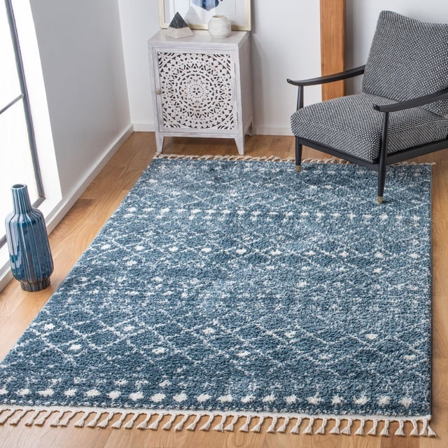 Safavieh Berber Fringe Robynne Tapis Shag Aztèque Bleu avec Motifs Tribaux Aztèques Blancs et Poil de 1,25 Po, avec Bords à Franges, Posé sur un Plancher de Bois Franc à Côté d'Un Fauteuil à Motif Gris et d'Une Armoire en Bois Sculptée Blanche, Accentué d'Un vase Bleu Près d'Une Fenêtre.
