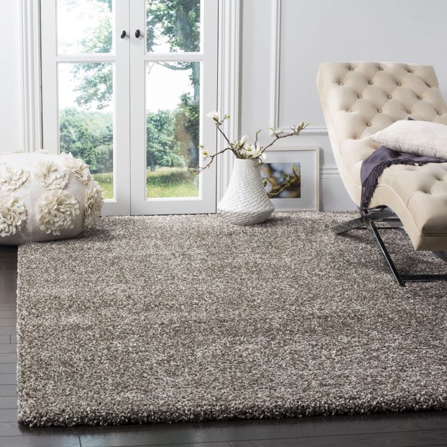 Safavieh Milan Harlow Tapis Shag Uni Gris avec un Poil de 2 Pouces d'Épaisseur, Exposé sur des Planchers de Bois Franc Sombres dans une Pièce Lumineuse. le Tapis en Fil Synthétique Tissé Mécaniquement Est Stylé avec une chaise Longue Tuftée beige, un vase Blanc avec des branches Florissantes, et un pouf floral Blanc Près de Portes en Verre Montrant une Vue Extérieure Verte. Idéal pour les Aires de Vie Très Passantes.