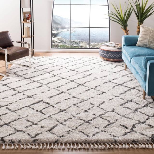 Safavieh Moroccan Fringe Tapis Shag Géométrique Dorien pour Salon Ensoleillé avec Grande Fenêtre Montrant une Côte Rocheuse, Présentant un Tapis Shag en Fil Synthétique Blanc Pelucheux avec Motif Géométrique Noir en Losanges et Bords à Franges, Assorti à un sofa Bleu, une Chaise en Cuir Brun et un pouf à Motifs, Convient à une Déco Moderne Ou Campagnarde Chic.