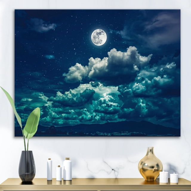 Designart Full Moon In Cloudy Night Sky II Canvas Wall Art featuring a gallery-wrapped cotton canvas print of a luminous full moon above thick, illuminated clouds in a deep blue starry night sky, stretched over a wood frame, displayed above a modern console with decorative vases and candles, arriving ready to hang and suitable for enhancing nautical or coastal home décor.