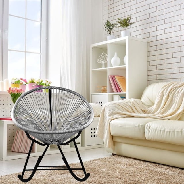 Costa Rocker Lounge Chair in grey with oval wire seat and black metal rocking frame, placed in a bright indoor living room featuring a cream leather sofa with beige throw, white shelving with plants and decor, and a large window with sheer curtains.