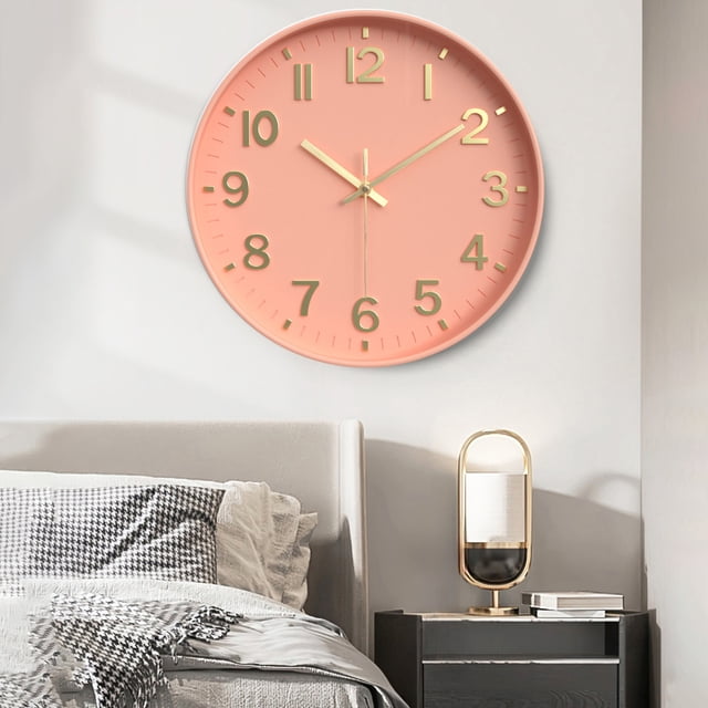 EGNMCR Wall Clock Silent Wall Clocks Battery Operated 12 Inch Modern Non Ticking Analog Clock. This decorative wall clock features a pink plastic frame with gold-tone hands and numerals, measuring 30x30x8cm (11.8x11.8x3.1in). It is displayed in a bedroom setting, mounted above a bed with a nightstand and lamp nearby. Ideal for kitchens, offices, bathrooms, and bedrooms.