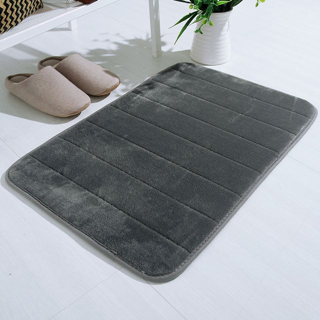 Agiferg Absorbent Soft Memory Foam Mat in grey, measuring 40x60cm (17x24 inches). This decorative bath rug is made from coral fleece with a memory foam polyurethane backing, offering a soft and absorbent surface. Suitable for use in bathrooms, bedrooms, or as a floor shower rug. The mat features a non-slip design and is machine washable for easy maintenance. Displayed in a bright, minimalist room with slippers and a potted plant nearby.