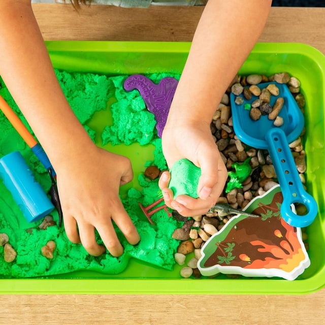 Made By Me! Explore + Create Dinosaur Sensory Bin with green moldable sand, natural pebbles, blue shovel, purple dinosaur mold, foam volcano cutout, and dinosaur figurines in a green plastic bin, suitable for sensory play and tactile learning for children aged 3 and up.