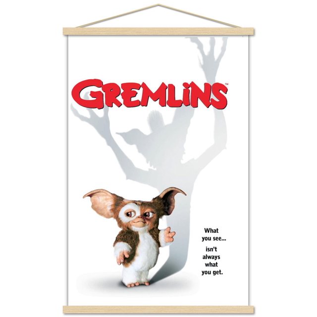 Gremlins - One Sheet Wall Poster, 22.375" x 34" featuring a small Mogwai character with brown and white fur and large ears standing on a white background. Its shadow behind it forms a large gremlin with outstretched claws. Bold red 'GREMLINS' text is at the top, with the tagline 'What you see... isn't always what you get.' on the right. The poster is framed with light wood hanging strips.