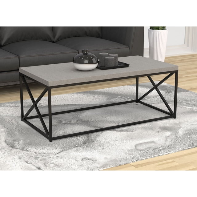 Safdie & Co. Coffee Table 48"L Cement with a thick rectangular cement-grain finish top and minimalist black metal frame featuring geometric cross supports, placed on a textured light grey rug in front of a dark grey sofa in a modern living room. Its sleek design is also suitable for use as an accent table.